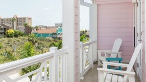 A balcony with white railings, two Adirondack chairs, and a view of a residential area with houses and greenery. - Luxury Coastal Retreat​​​​​​​ • Heated Pool & Hot Tub • 3 Min Walk to Beach (South Padre Island)