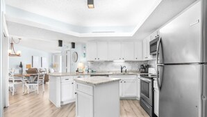 A modern kitchen with white cabinetry, a central island, stainless steel appliances, and a dining area with a wooden table and chairs. - Totally Beachin - Condo with Stunning Ocean Views (Orange Beach)