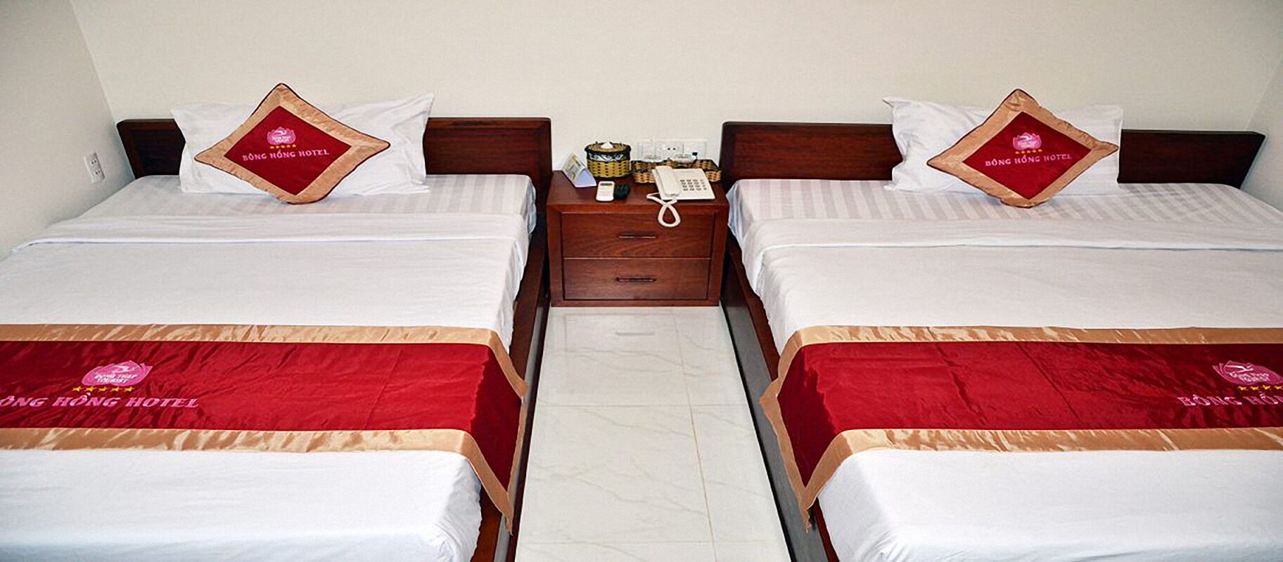 Two identical twin beds with red and white bedding, a wooden nightstand with a telephone and a basket between them, and a white wall in the background.