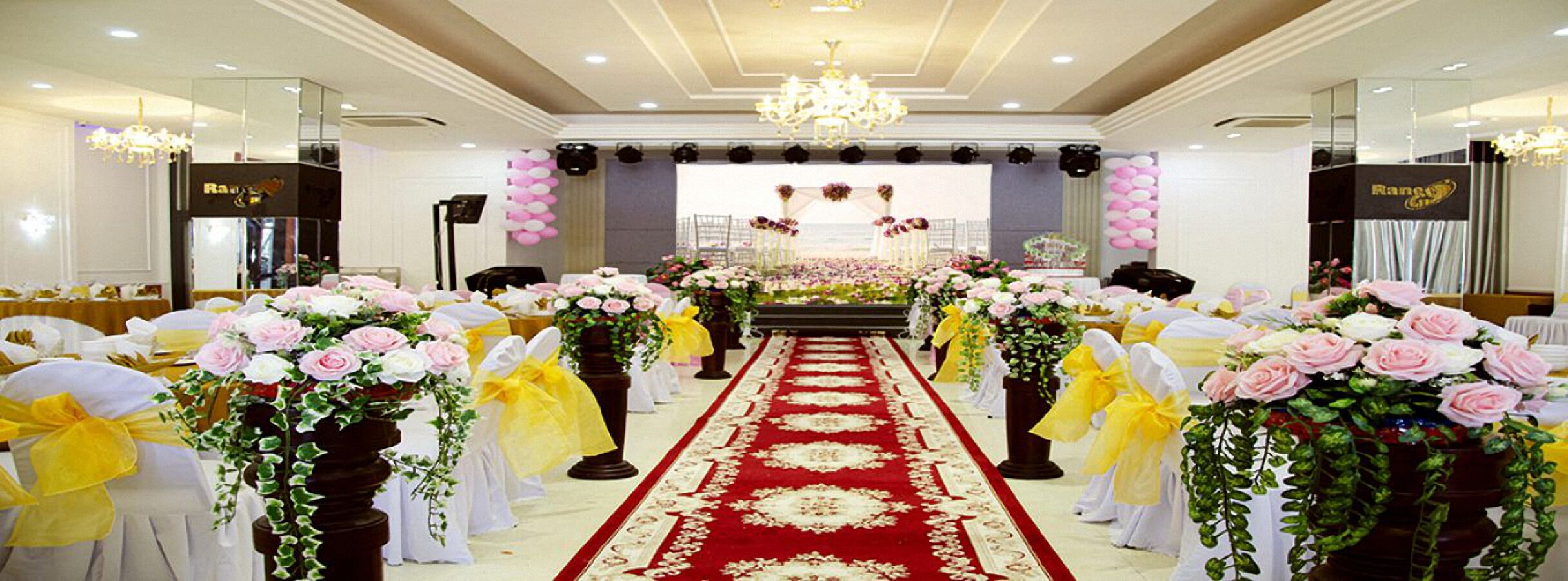 A wedding ceremony setup with a red carpet, floral arrangements, and a stage with a backdrop.