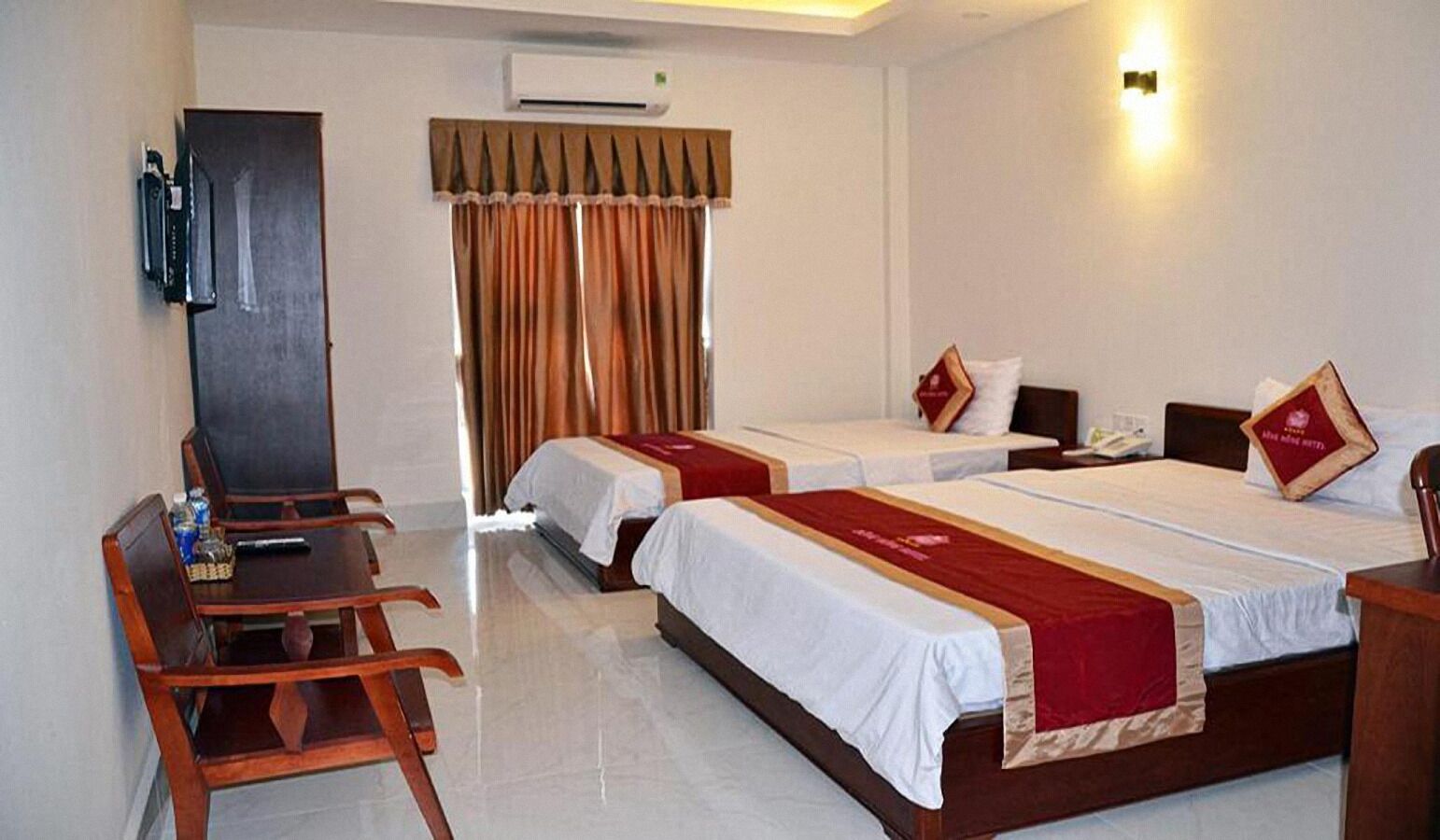 Une chambre d’hôtel avec deux lits simples, un bureau avec des chaises, un minibar et une grande fenêtre avec des rideaux.