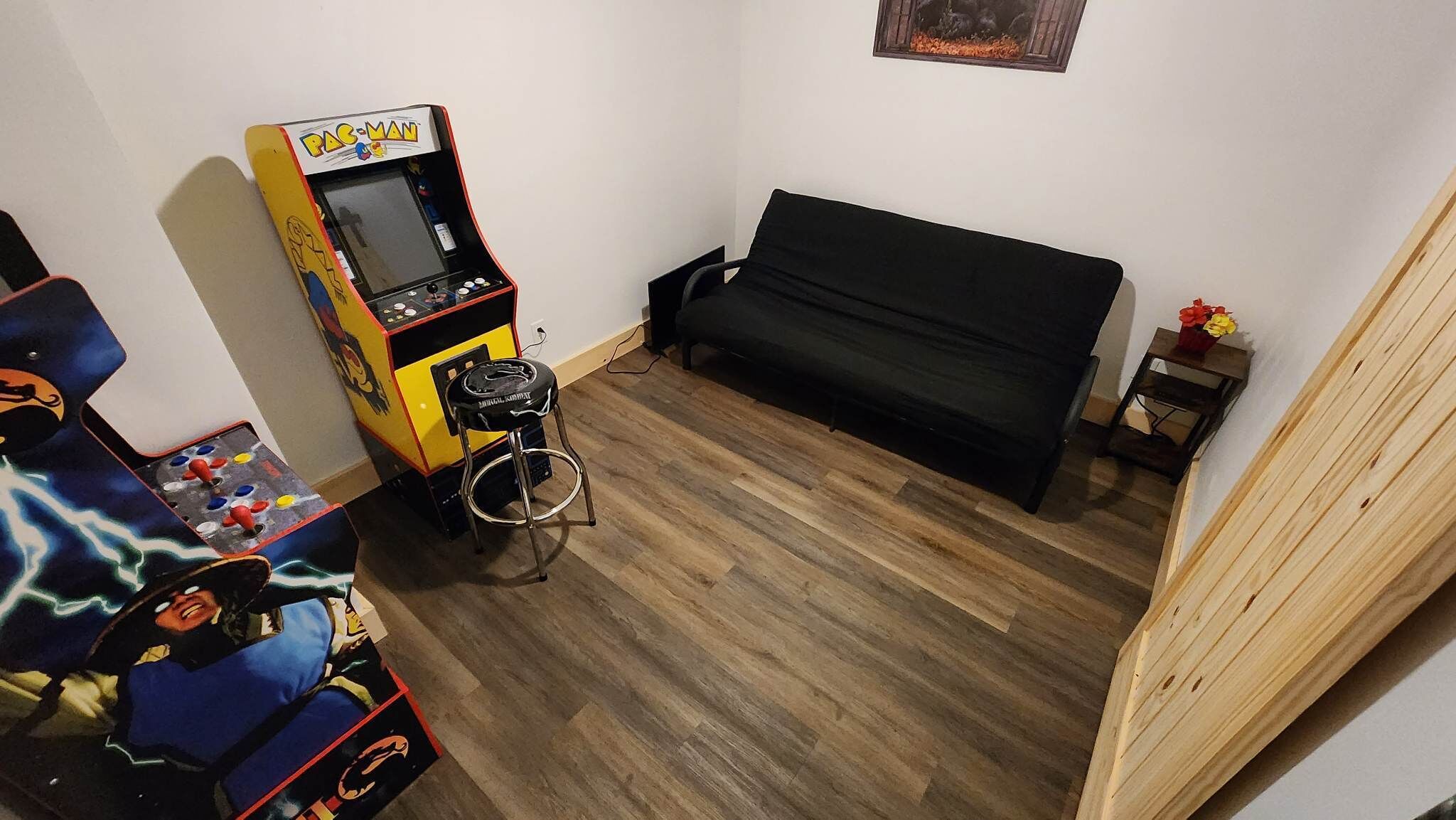Een kamer met een houten vloer, een zwarte bank, een Pac-Man spelcomputer en een kleine tafel met een bloevas.