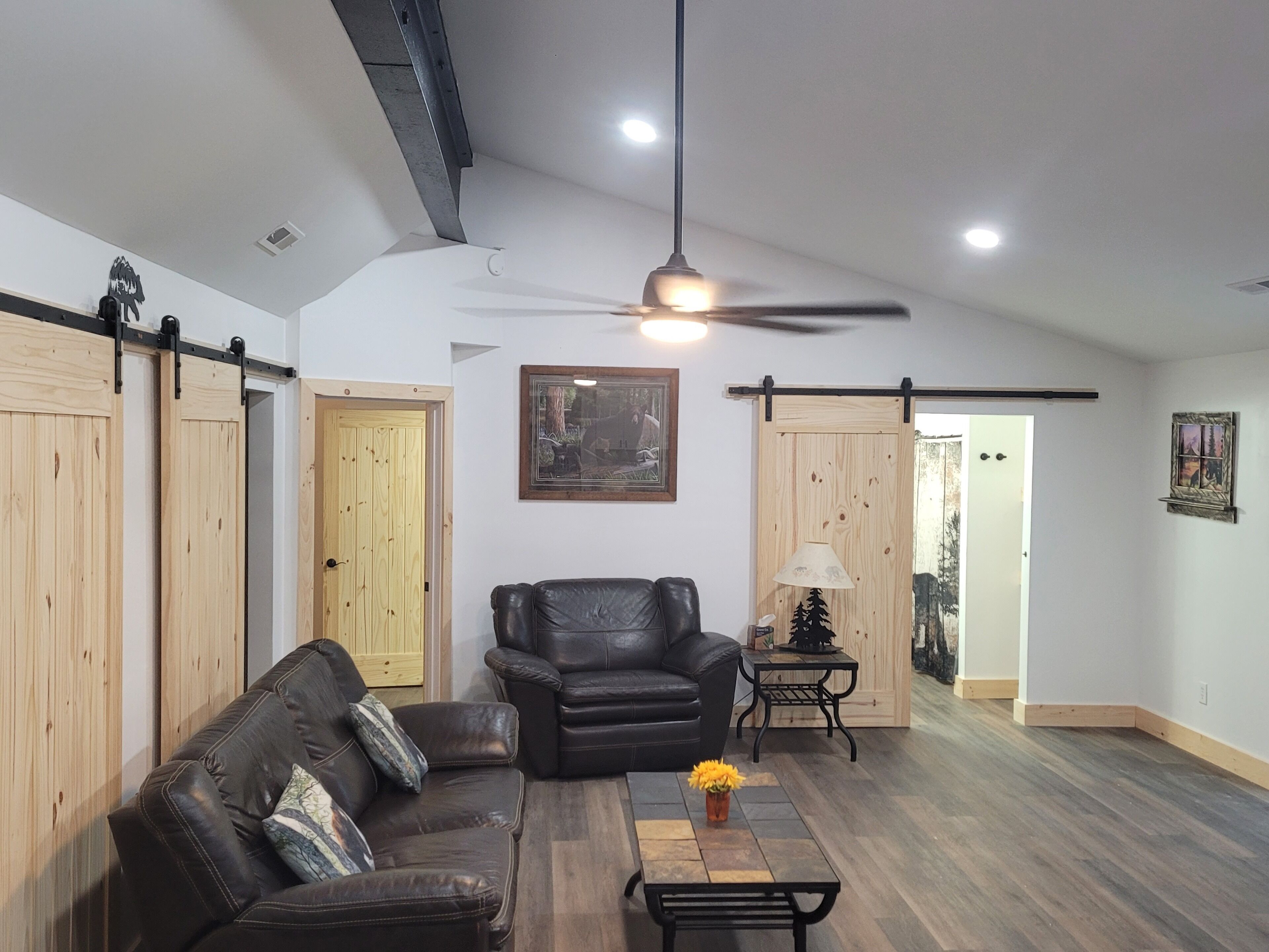 Una sala de estar con un sofá de cuero, un sillón negro, una mesa de centro con flores y un ventilador de techo.