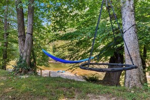 A hammock swing suspended between trees in a wooded area with a dirt path nearby.