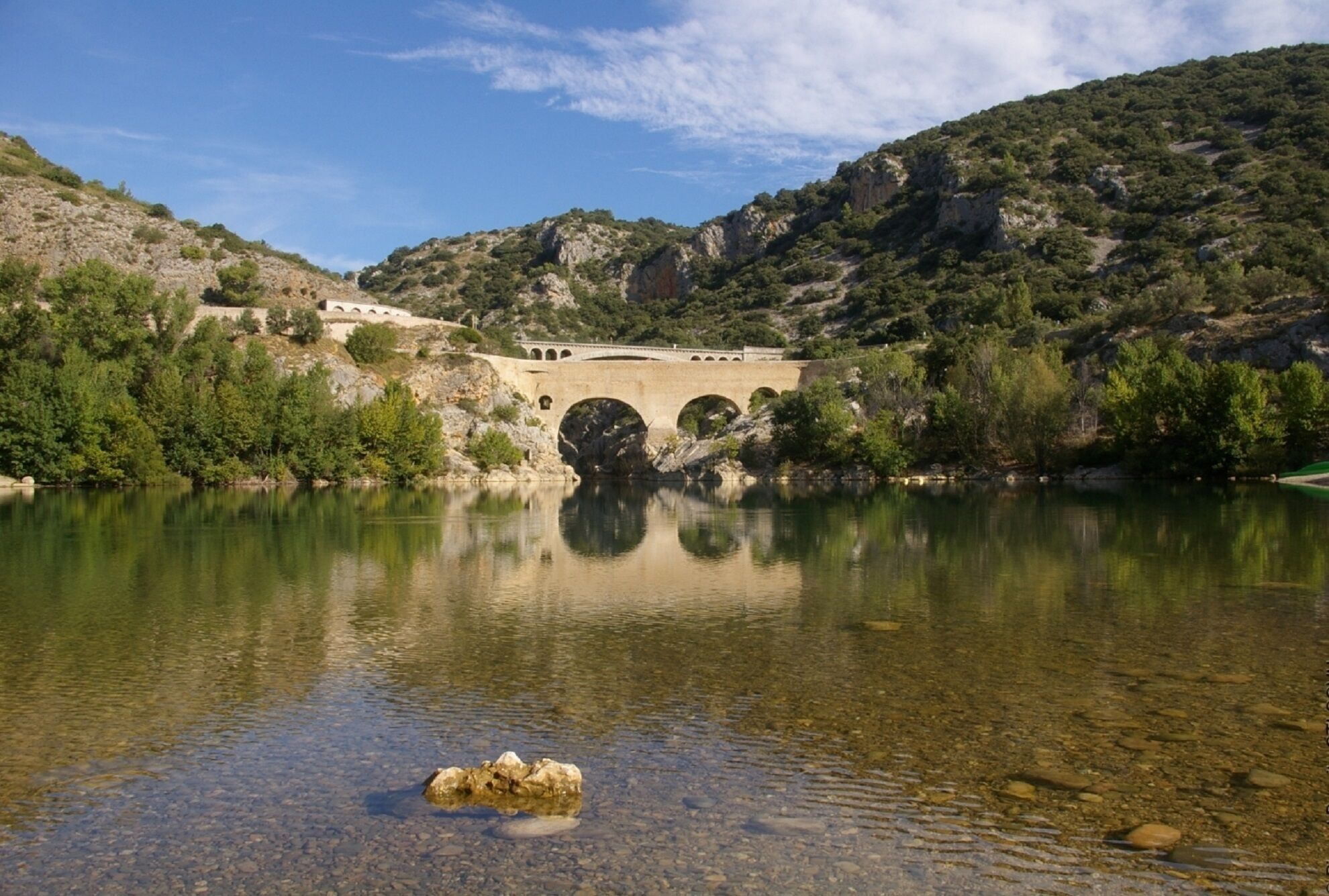 Un pont de pierre enjambe un lac aux eaux claires, avec des collines rocheuses en arrière-plan.