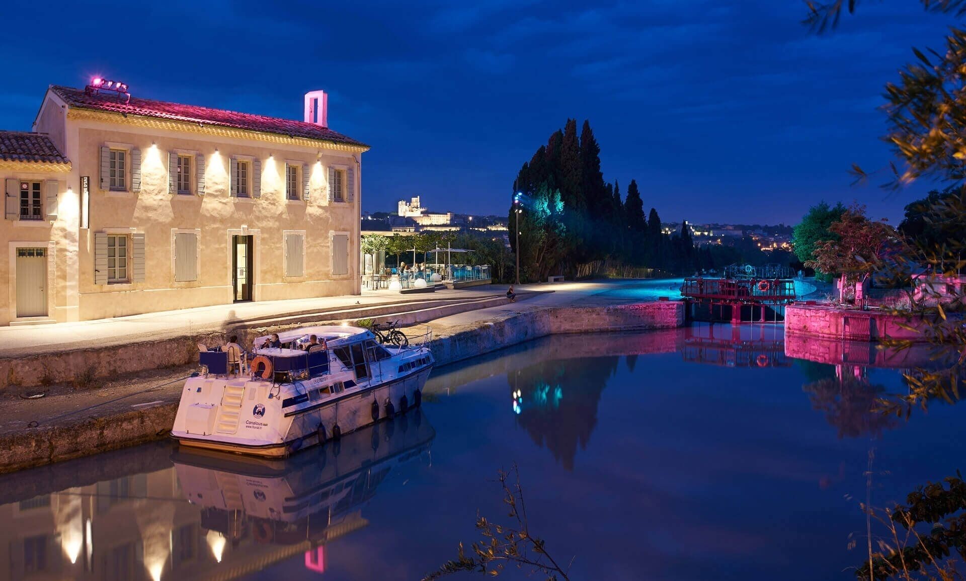 Un río con una barca amarrada en un muelle, un edificio con tejado rojo y un paisaje urbano al fondo.