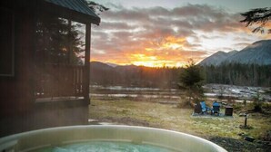 A hot tub with a view of a sunset over mountains and a wooden cabin.