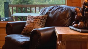 A cozy cabin interior with a leather chair, a wooden table, and a view of a forested mountain.