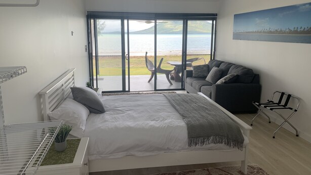 A bedroom with a bed, a sofa, a chair, and a large sliding glass door leading to a balcony with a view of the sea. - Sinclair Bay Resort (Unit 1) - Peaceful waterfront retreat on private estate (PROSERPINE)