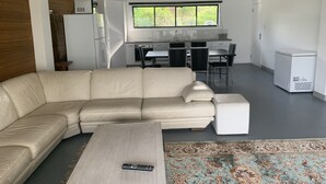 A modern living room with a beige sectional sofa, a wooden coffee table, and a remote control on it. There is a kitchen area with a dining table and chairs, a refrigerator, and a window with a view of greenery. - Sinclair Bay Resort (Unit 1) - Peaceful waterfront retreat on private estate (PROSERPINE)