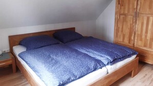 Un lit simple avec une tête de lit en bois et une couvre-lit bleue, dans une pièce dont le sol et l’armoire sont en bois.