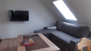 Un salon moderne avec un canapé gris, une table basse en bois sur laquelle sont posés le jeu de société Monopoly et d’autres jeux de société, un téléviseur à écran plat fixé au mur et une lucarne.