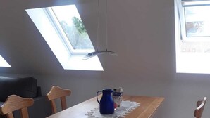 Un coin repas avec une table et des chaises en bois, un pichet bleu et une nappe en dentelle. Des lucarnes se trouvent au plafond.