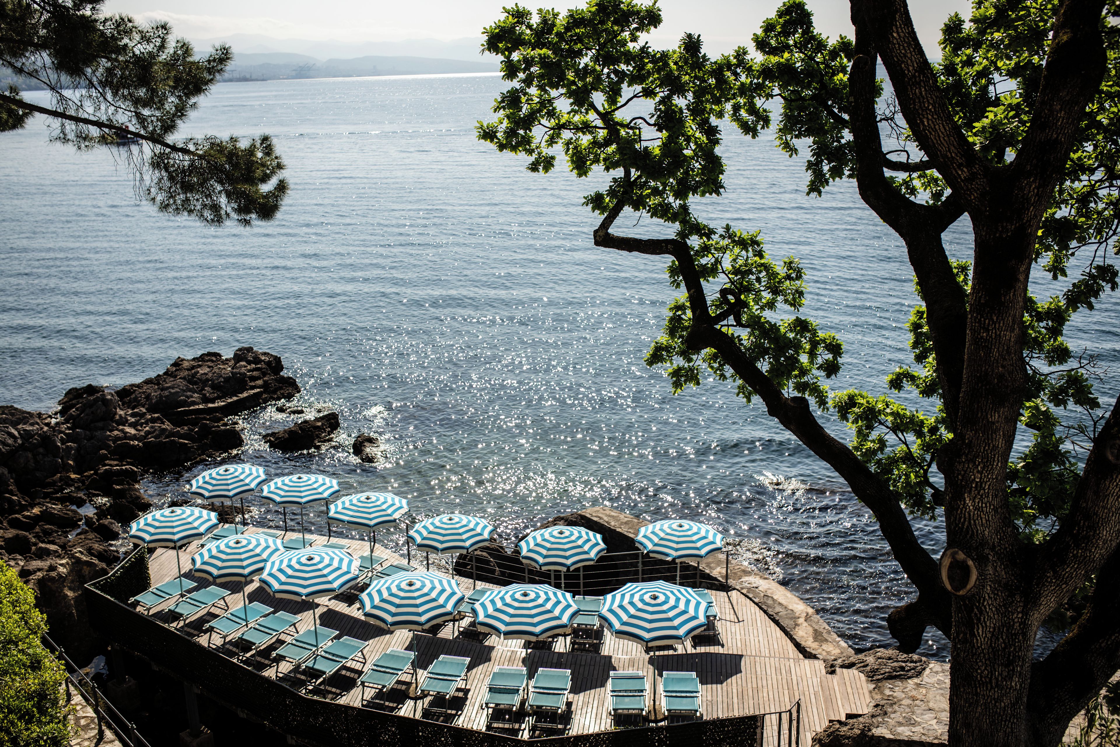 A seaside terrace with multiple sun loungers and striped umbrellas, overlooking a rocky coastline and the sea.