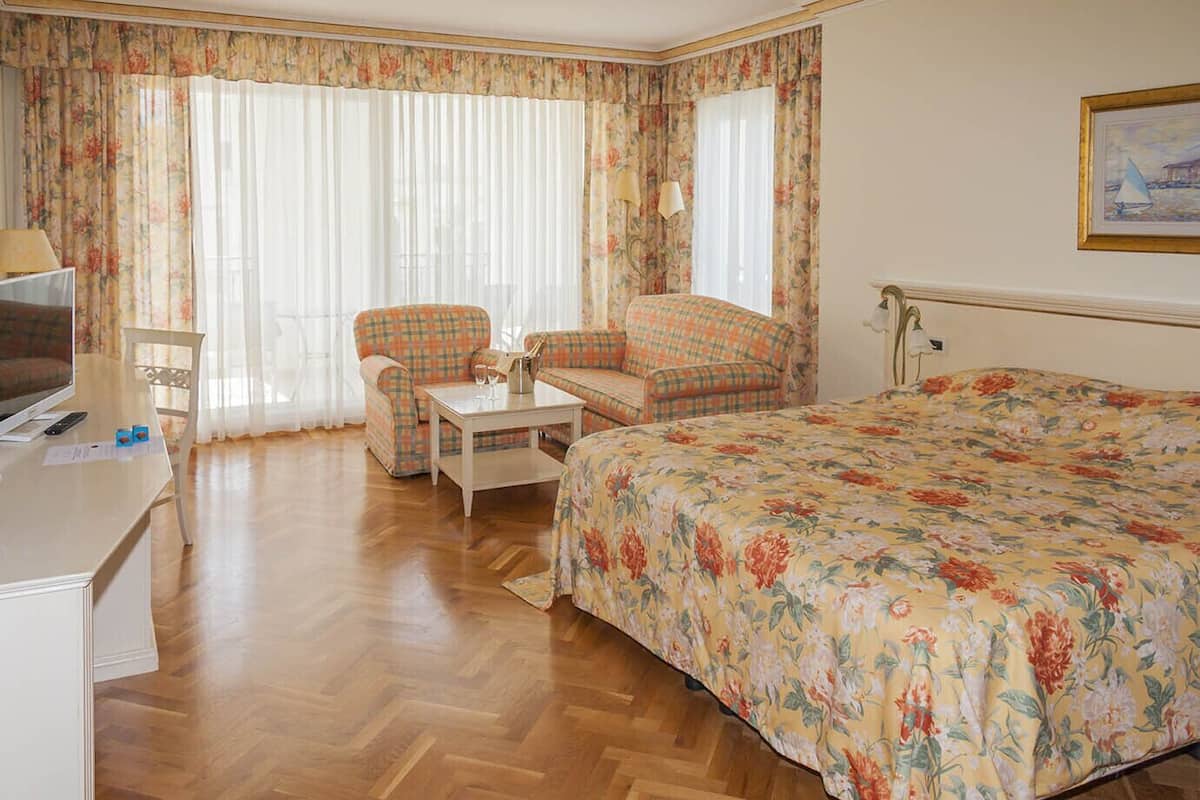 Habitación de hotel con una cama grande, dos sillones, una mesita, un escritorio con televisor y una ventana con cortinas.