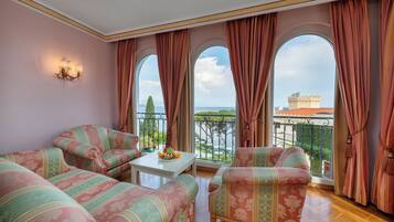 A room with a sofa set, a small table with fruit, and a view of a castle through large windows.