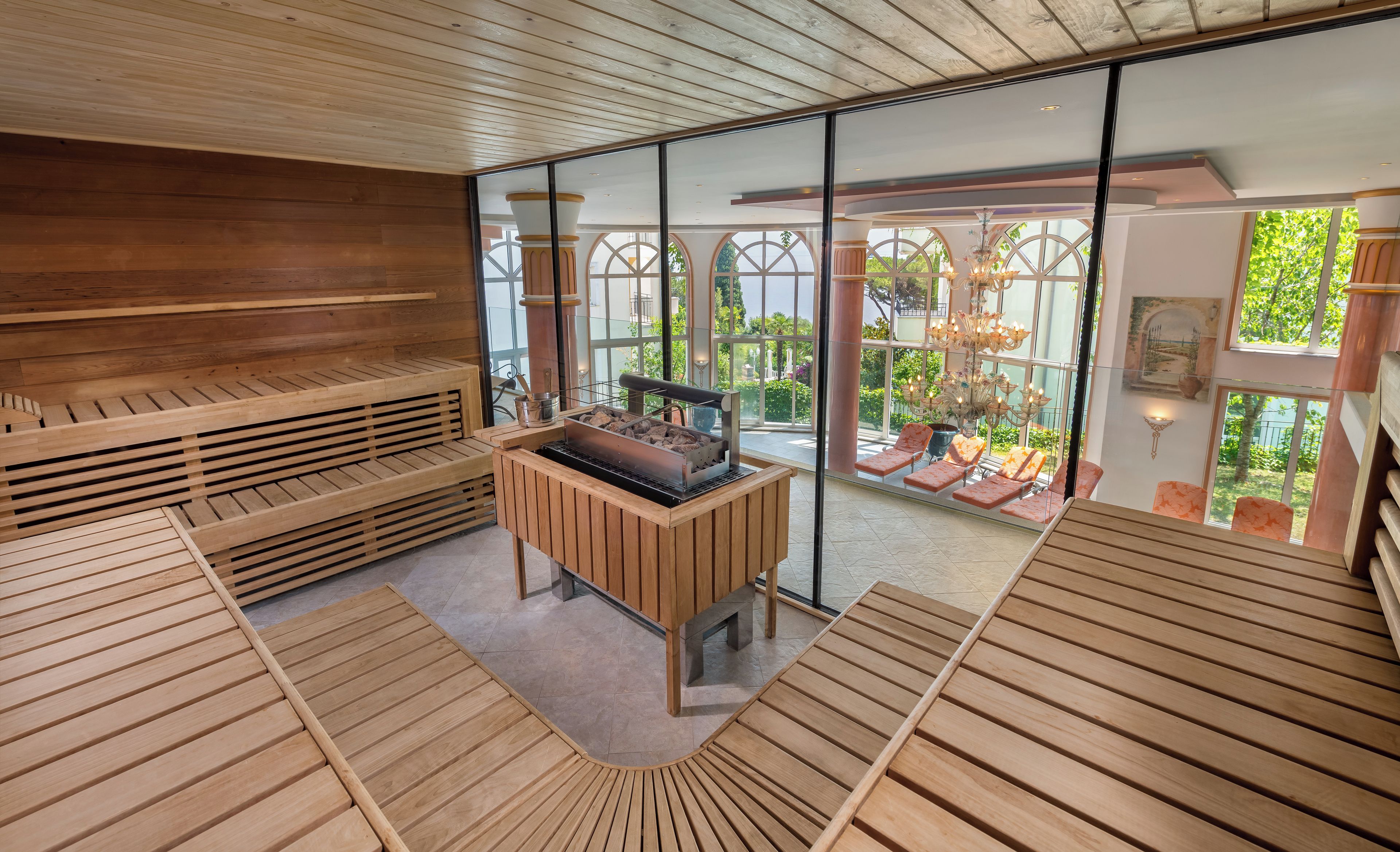 A sauna with wooden benches and a central heating element, large windows with arched designs, and a view of greenery outside.