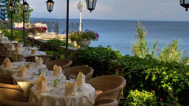 Un espace de restauration en plein air, avec des chaises et des tables en osier disposées pour un repas, offrant une vue sur une étendue d’eau.