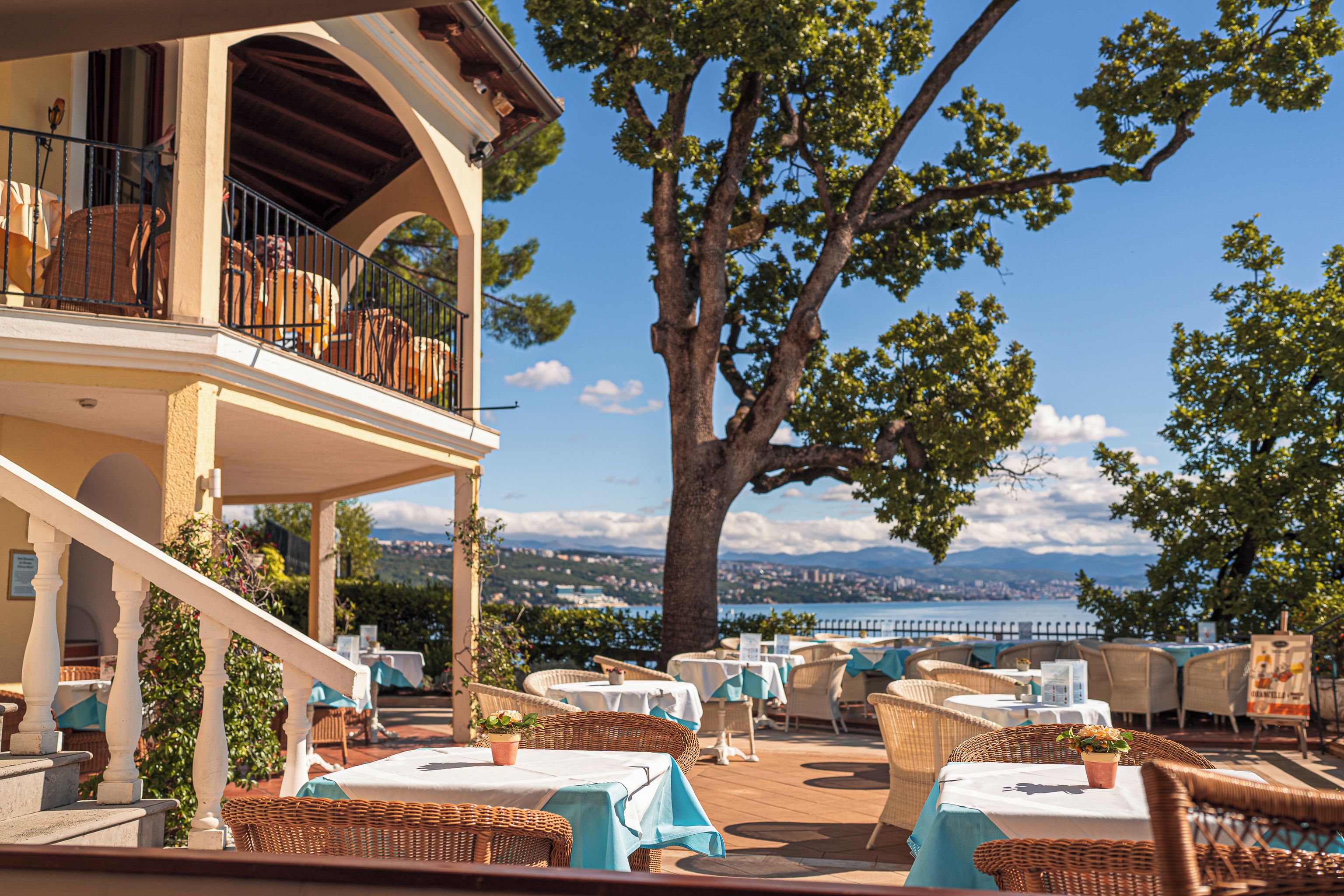 An outdoor seating area with wicker furniture and tables, overlooking a scenic view of a body of water and distant mountains.