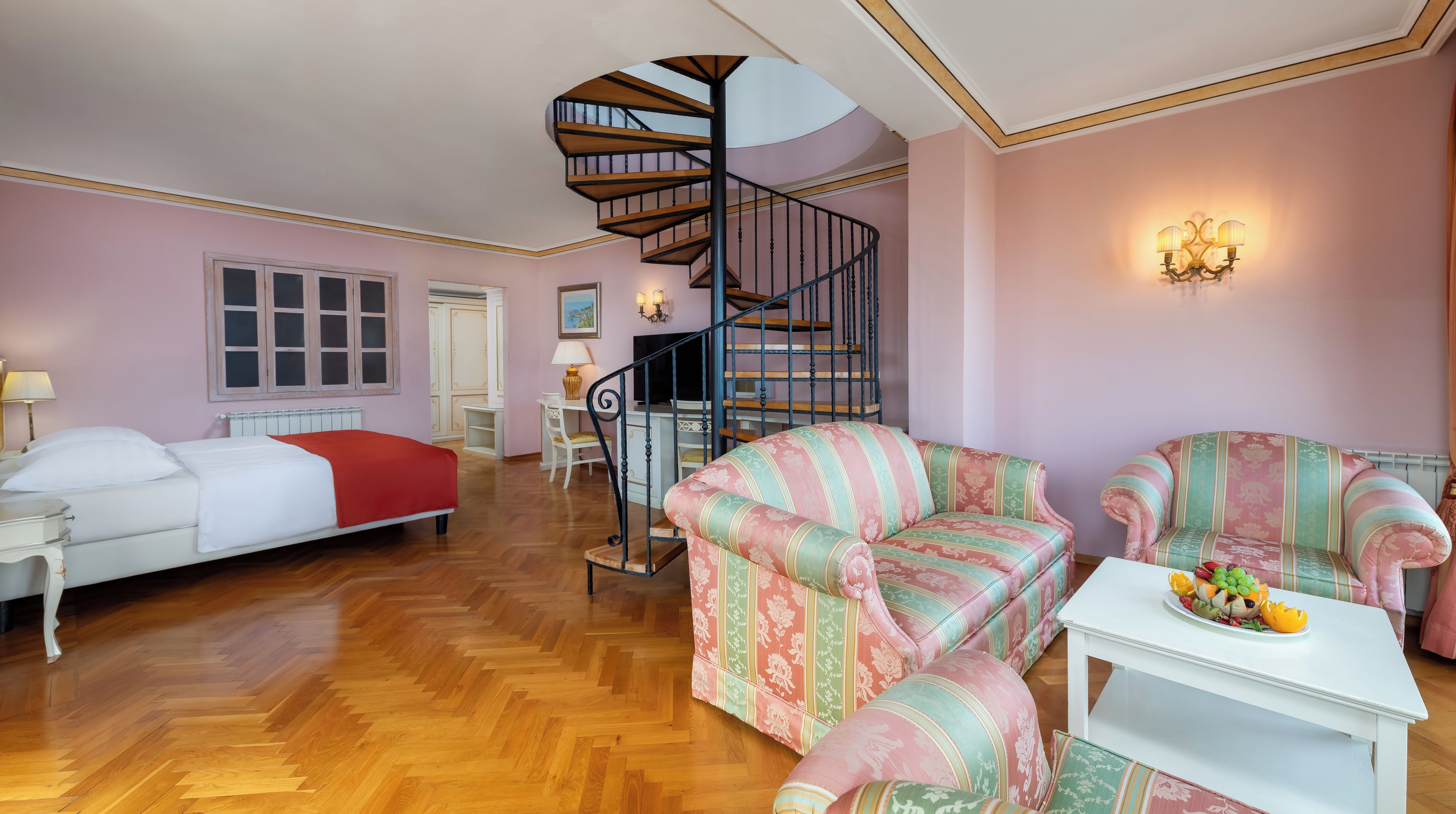 A hotel room with a bed, a sofa, a chair, a small table with fruit, a TV, a lamp, a window, and a spiral staircase.