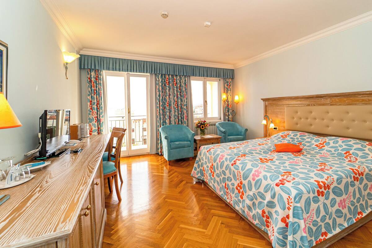 Habitación de hotel con un escritorio de madera, dos sillones, una cama con cubrecama floral y un balcón con cortinas.