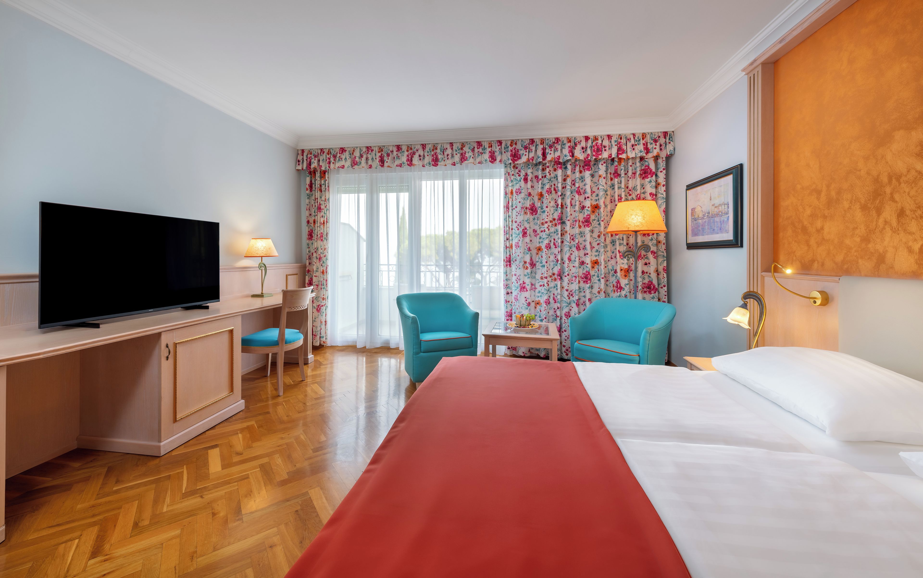 Habitación de hotel con cama, escritorio con televisor, dos sillas, mesita de noche con lámpara y ventana con cortinas florales.