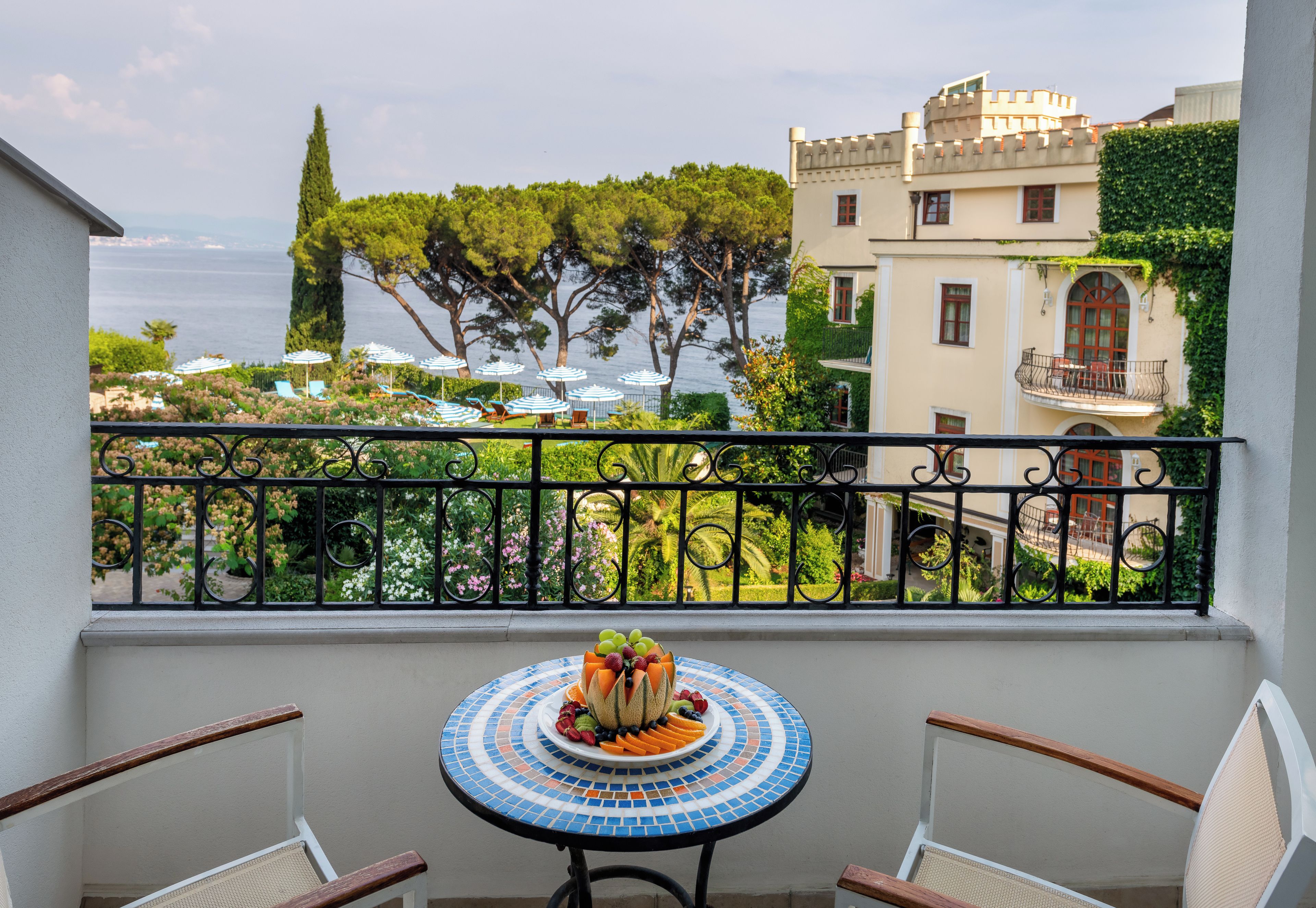 A balcony with a table set for a meal, overlooking a garden with trees and a view of the sea.