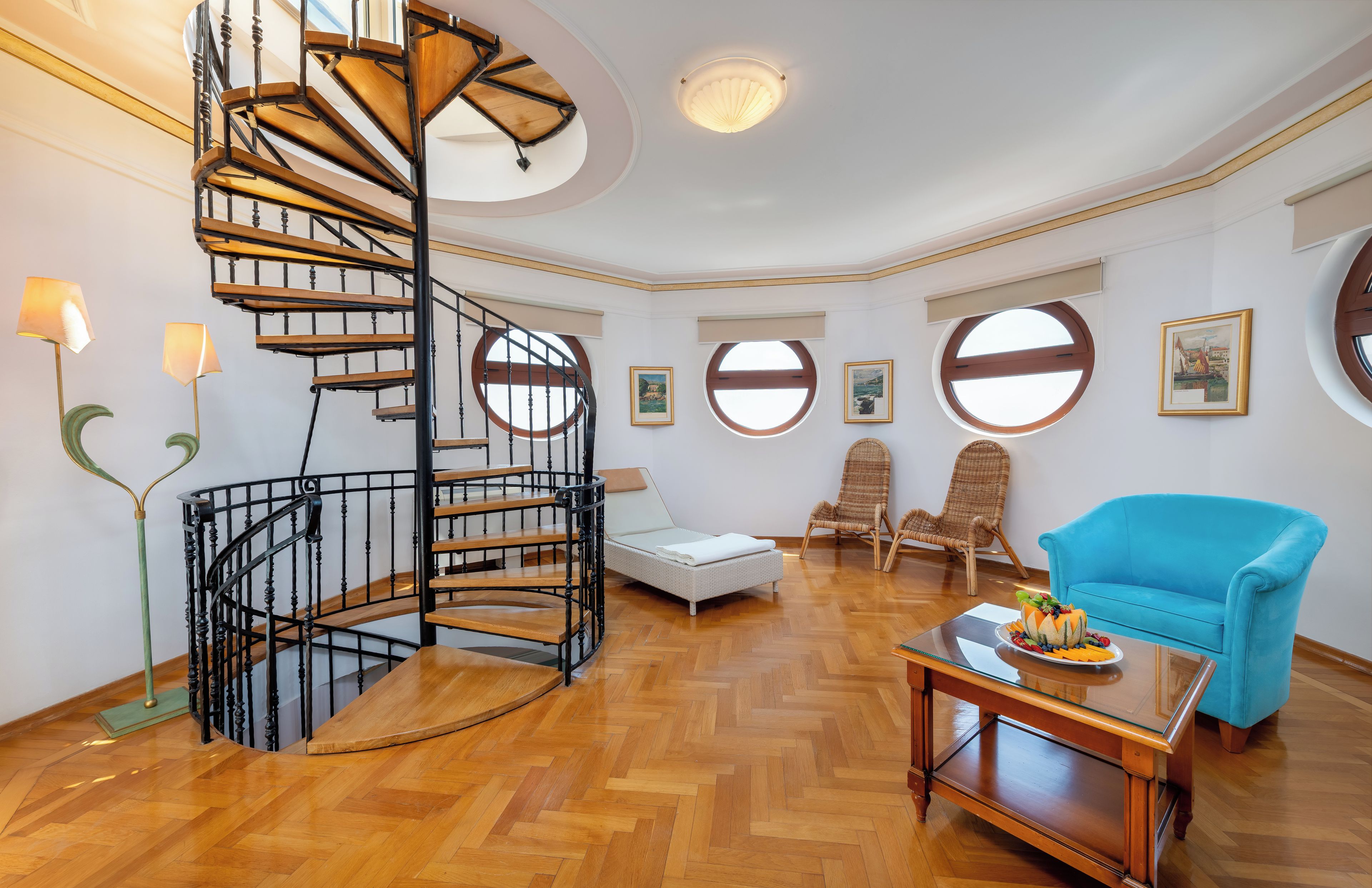 A spacious room with a spiral staircase, round windows, a blue sofa, and a dining table with fruit.