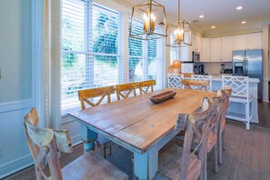 Un coin repas avec une table et des chaises en bois, une cuisine à l’arrière-plan et de grandes fenêtres équipées de stores.
