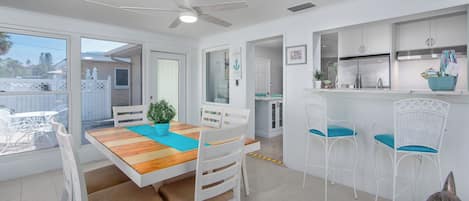 A bright kitchen with white cabinets, a stainless steel refrigerator, and a dining area with a wooden table and white chairs.