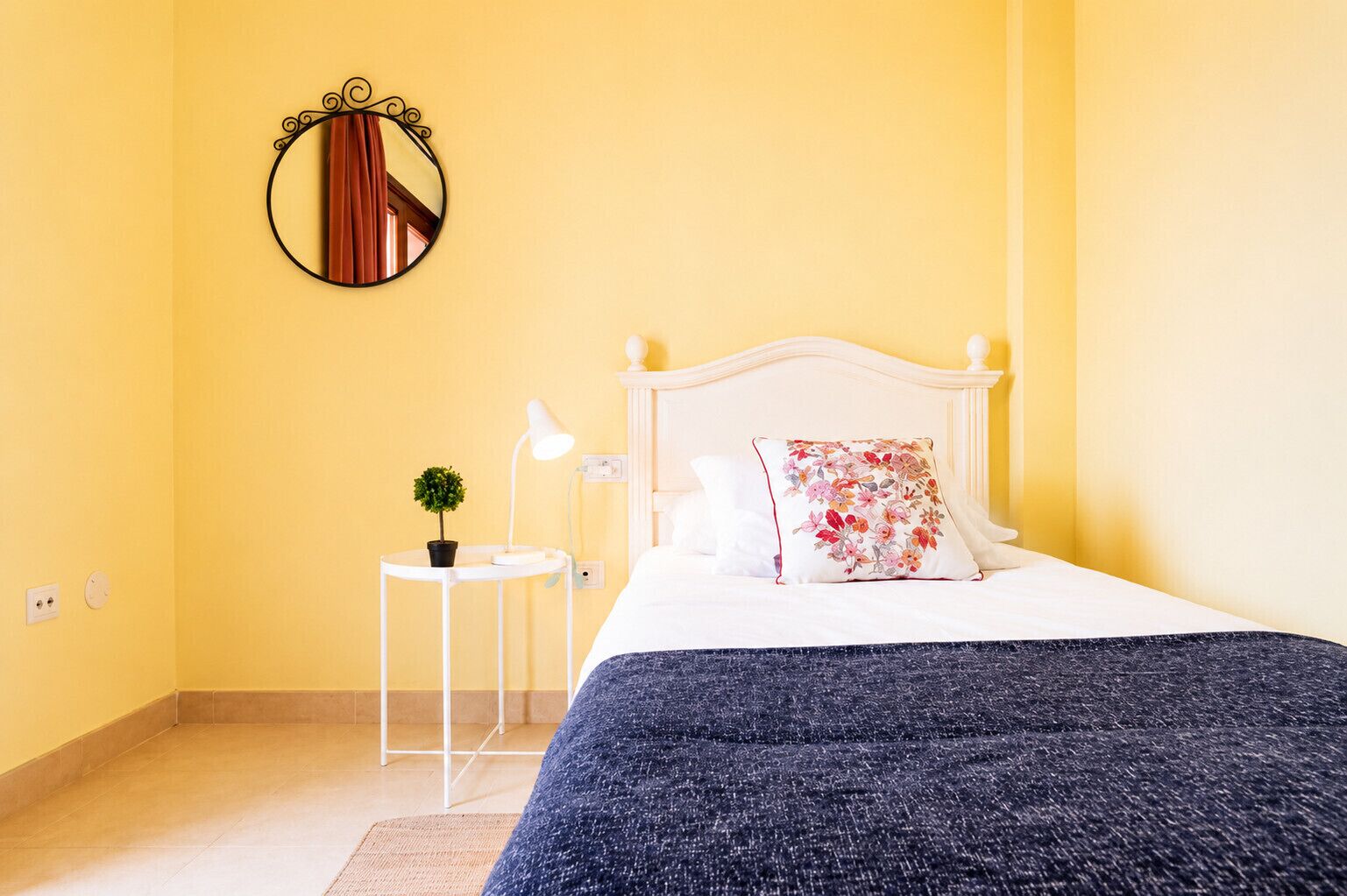 A bedroom with a bed, a bedside table with a lamp, a small plant, and a mirror on the wall.