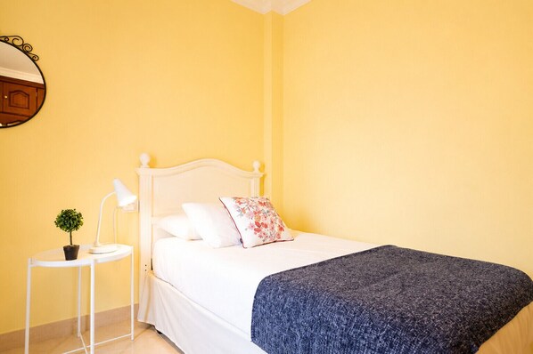 Ein Bett mit weißer Bettwäsche, ein kleiner Nachttisch mit Lampe und Pflanze sowie ein Spiegel an der Wand.