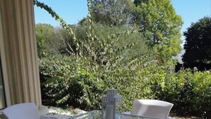 Patio esterno con tavolo in vetro e sedie bianche, circondato da piante e alberi.