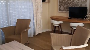 Un soggiorno con due poltrone, un tavolino, una TV su un supporto in legno e un'ampia finestra con tende.