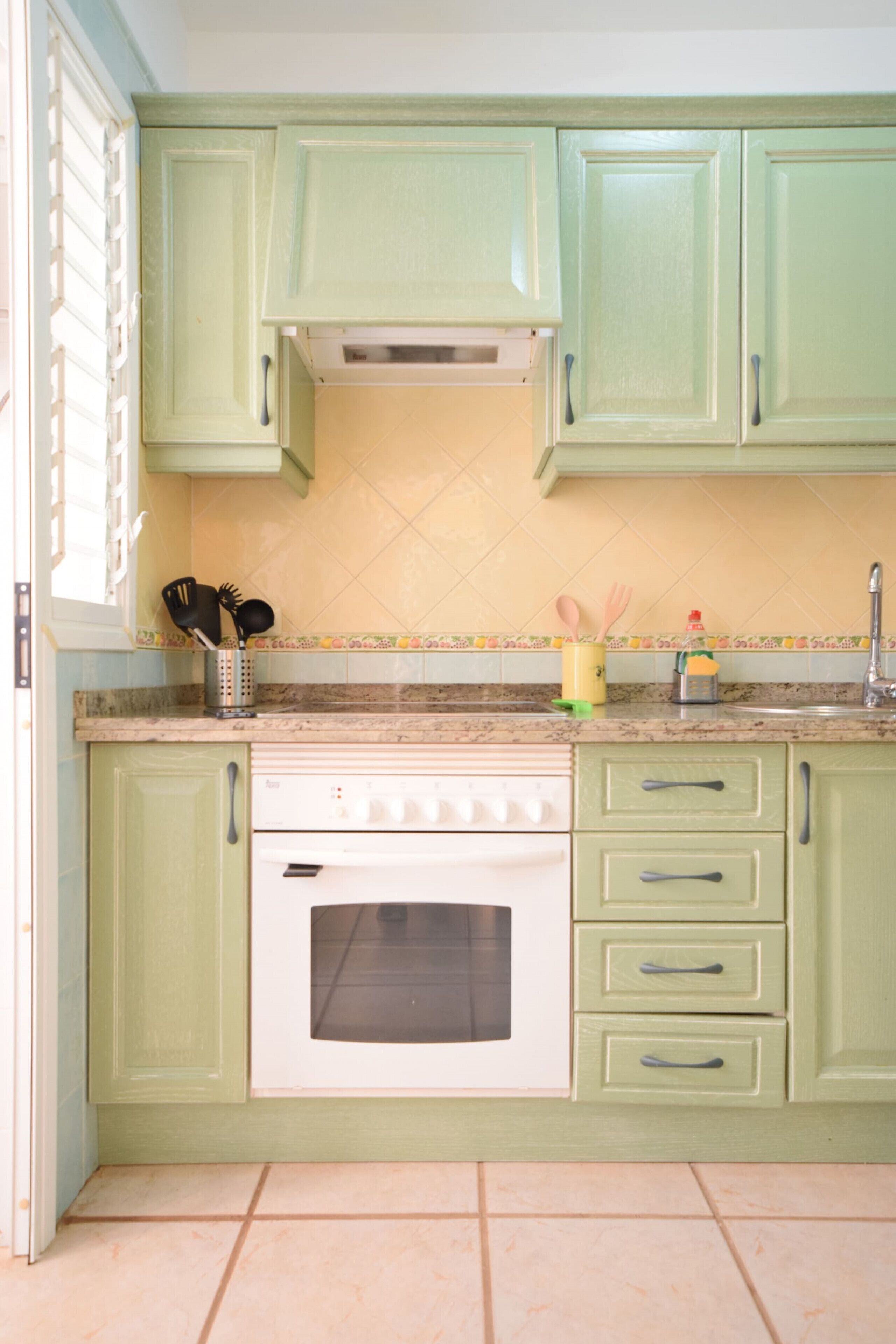 Een keuken met groene kastjes, een witte oven en een betegelde achterwand.