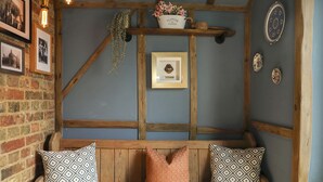 Un banco de madera con cojines estampados, un cuadro enmarcado y una estantería con objetos decorativos, todo frente a una pared de ladrillo.