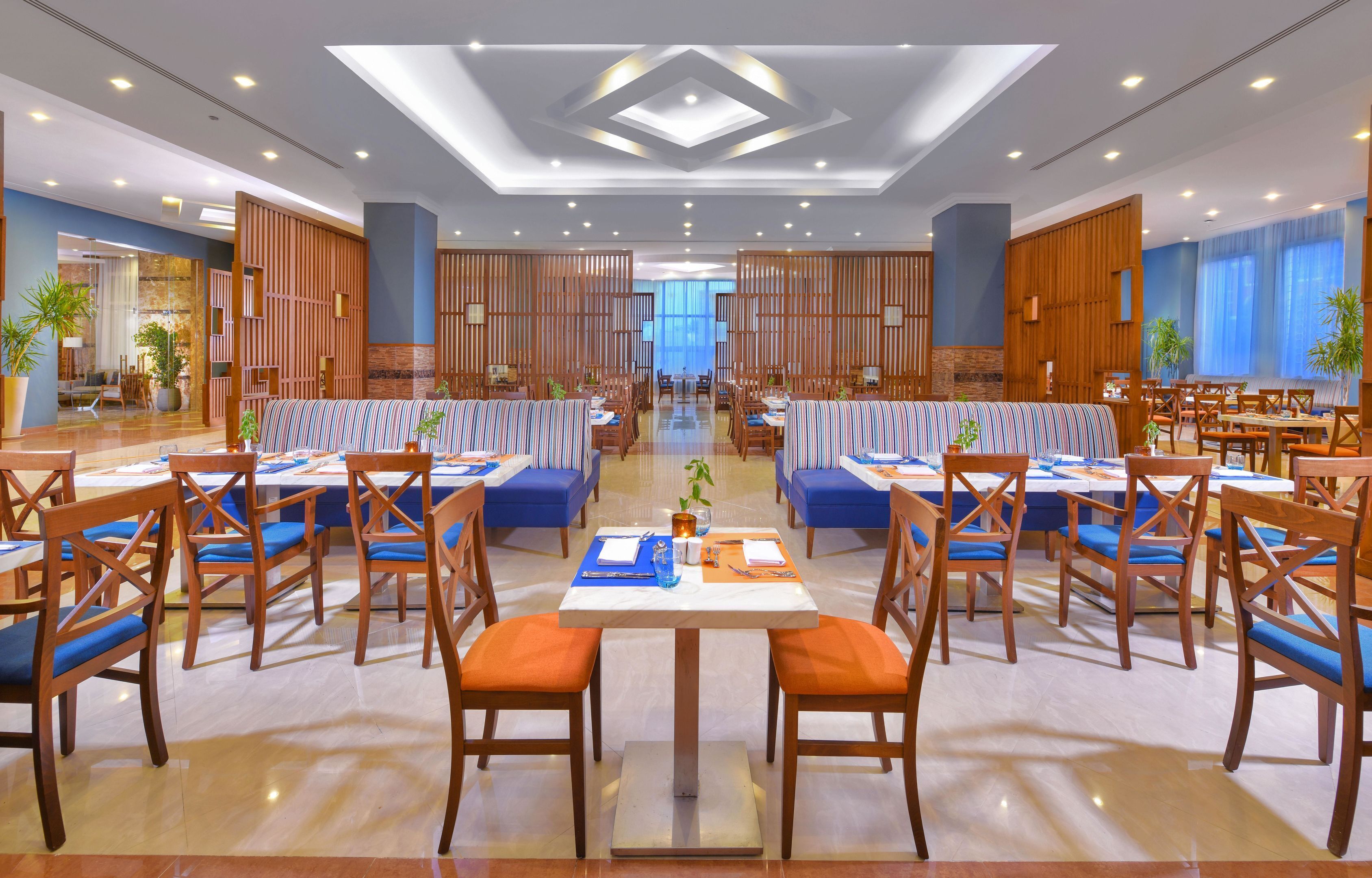 Un amplio vestíbulo de hotel con un área de descanso central, varias mesas de comedor y un diseño moderno en el techo.