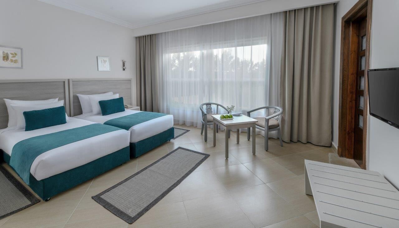 Ein Hotelzimmer mit zwei Betten, einem kleinen Tisch mit Stühlen, einem Fernseher und einem großen Fenster mit Vorhängen.