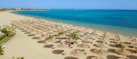 Una playa con numerosos sombrillas y sillas de descanso, un mar azul cristalino y una costa distante con edificios.