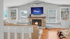 A living room with a fireplace, a ceiling fan, and a television displaying the Disney+ logo. - Modern Waterfront Lakehouse, with a private beach and a spacious boat dock. (Lake Placid)