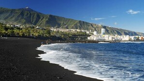 Una playa con arena negra, olas y un paisaje urbano con edificios y una montaña al fondo.