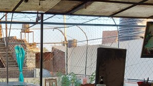 Una habitación con un amplio ventanal que da a una obra, una reja metálica y algo de vegetación.