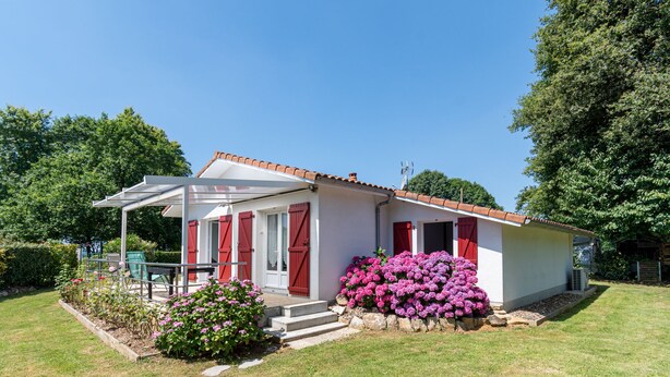 A small house with a covered porch, red shutters, and a garden with pink flowers.