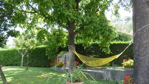 Una cadira de filferro penjada entre arbres a un jardí amb plantes en testos i una paret de pedra.