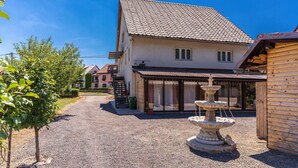 Ein Haus mit Kiesweg, einer Steinsäule mit Brunnen und einer Holzanbauten rechts.