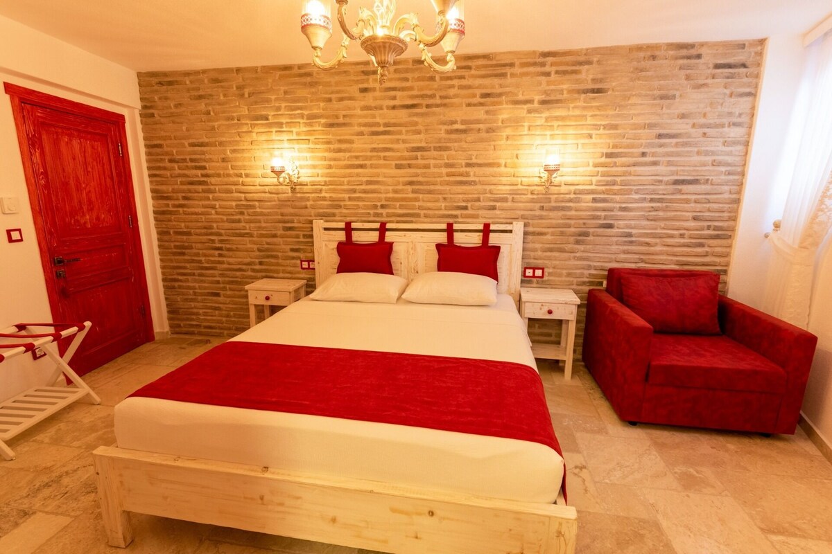 Une chambre avec un mur en briques, un lit en bois, deux tables de chevet, une chaise rouge et un lustre.