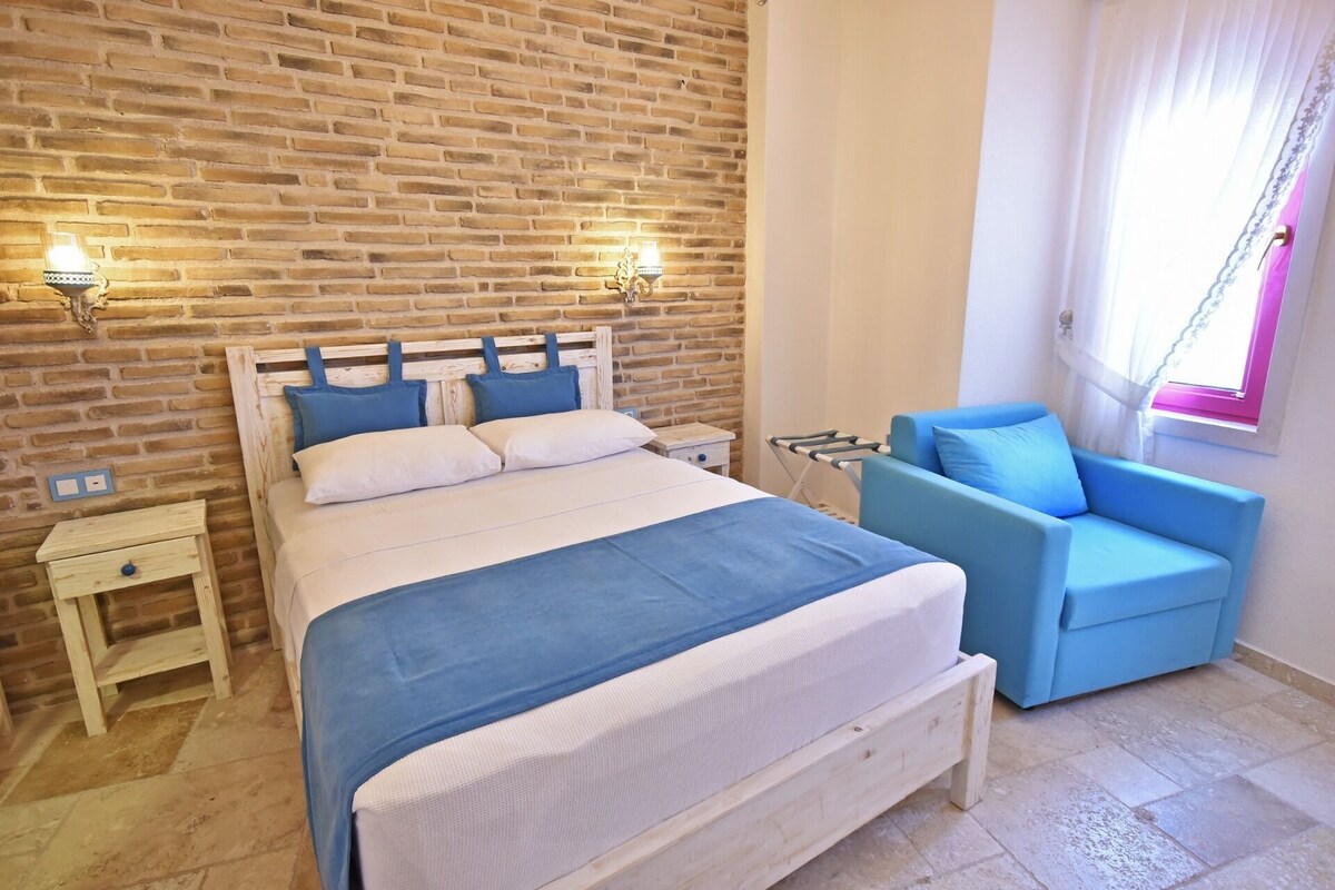 Une chambre avec un mur en briques, un lit avec une literie bleue, un fauteuil bleu et une fenêtre avec des rideaux.