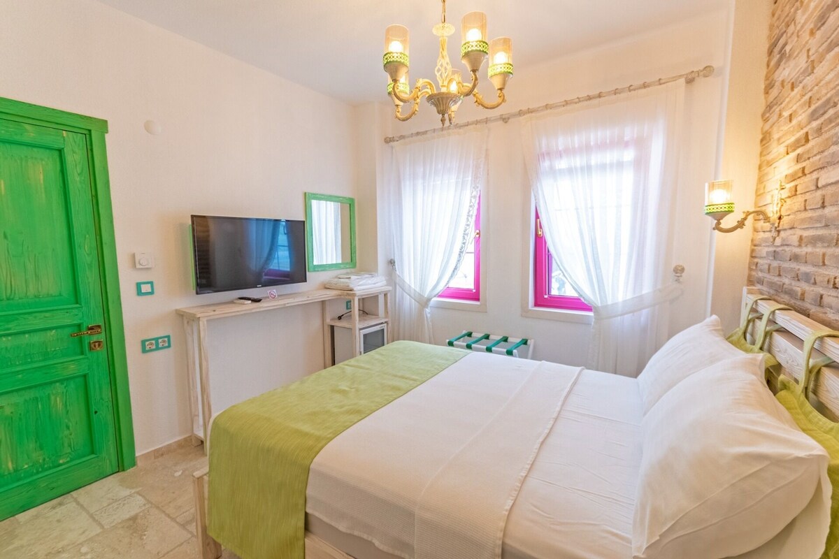 Une chambre avec une porte verte, un téléviseur posé sur un bureau, un lit avec une literie blanche et un lustre.
