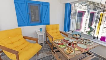Una habitación luminosa con contraventanas azules, una silla amarilla, una mesita con platos de comida y una ventana con vistas a edificios de colores.
