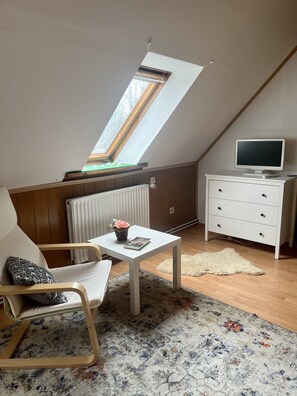 Un dormitorio acogedor en el ático, con techo inclinado, tragaluz, cómoda blanca, televisor, una silla, una mesita con un libro y un jarrón, y una alfombra estampada.